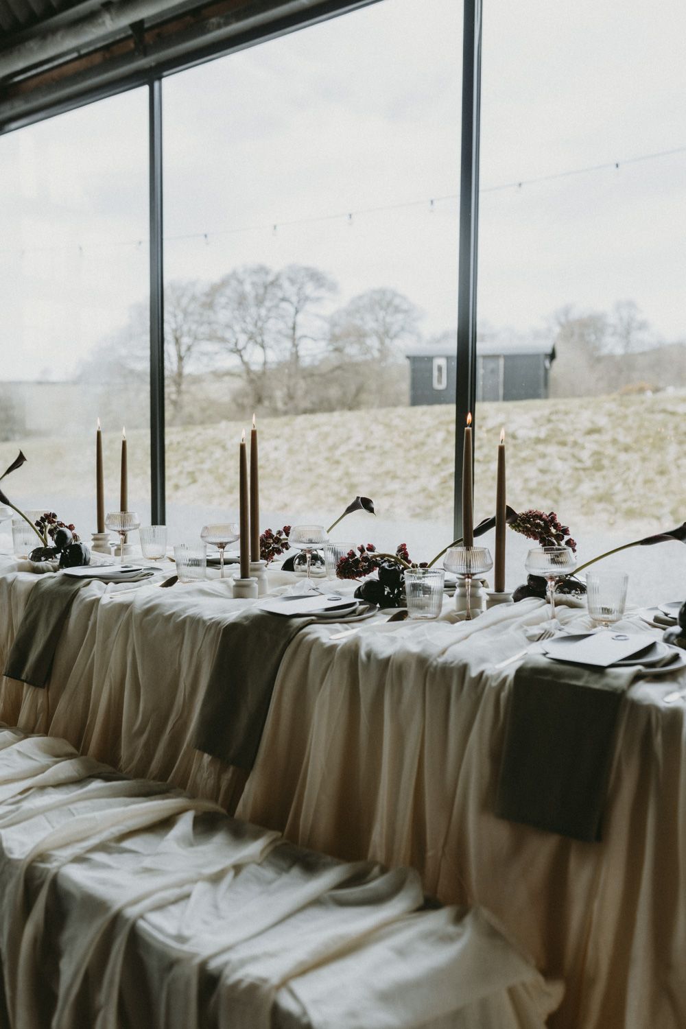 Elegant long dining table with tapered candles set against large windows overlooking winter landscape.