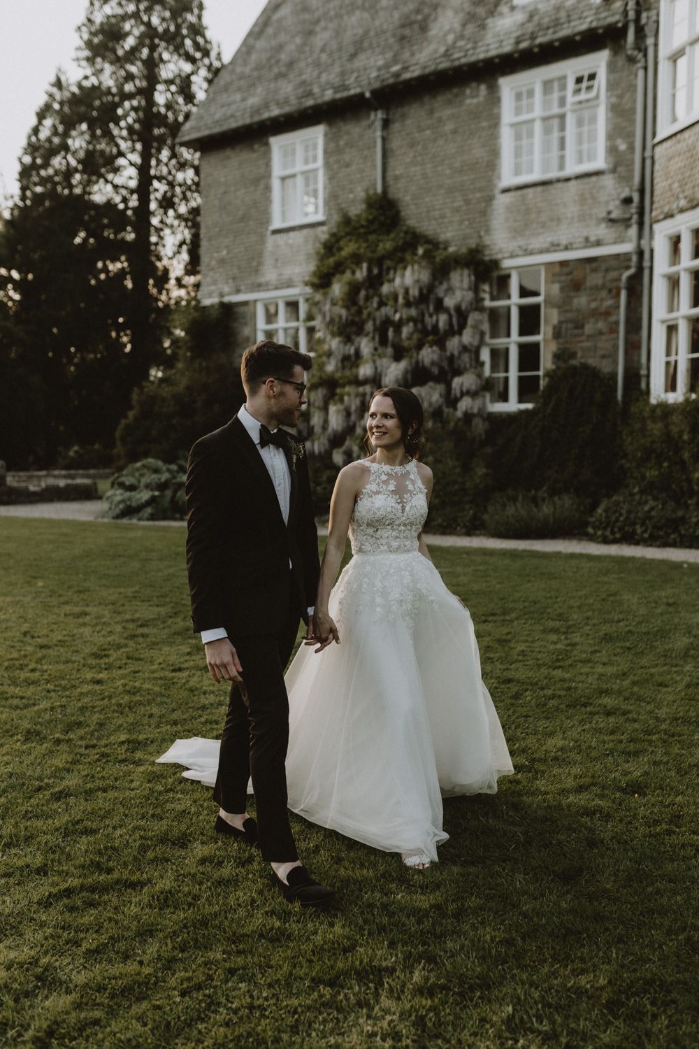 Newlyweds share a joyful moment on the lawn of a stone country house at dusk.