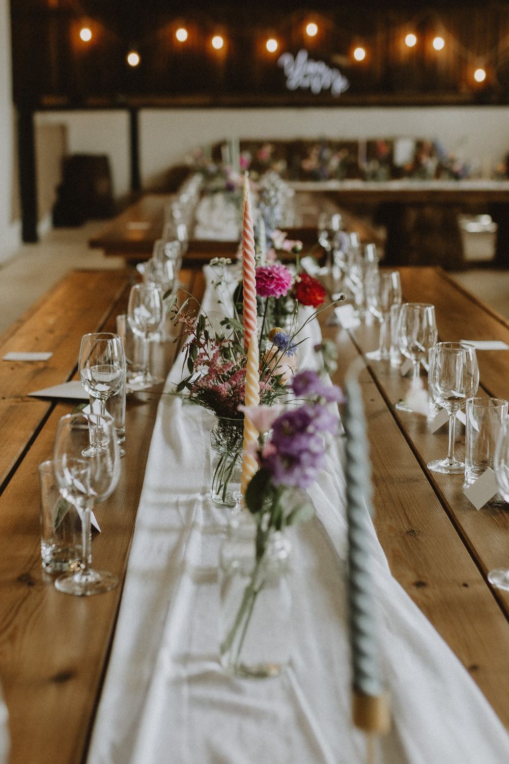 Wedding reception table settings with string lights, wine glasses, and purple floral centerpieces.