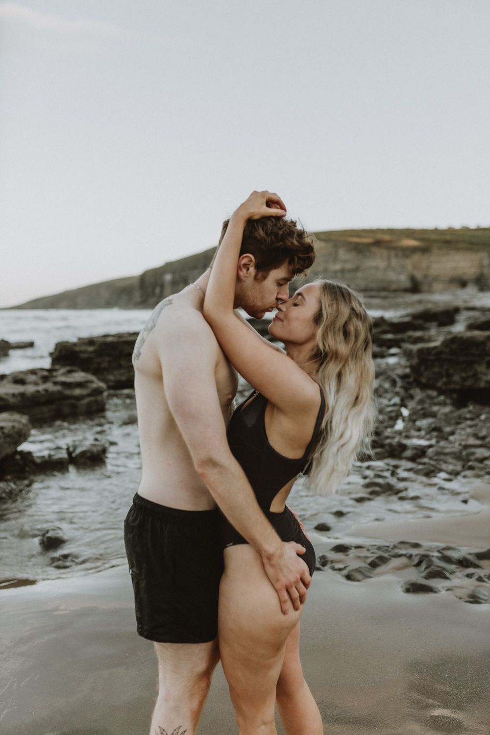 Intimate couple embrace on rocky beach during sunset photoshoot.