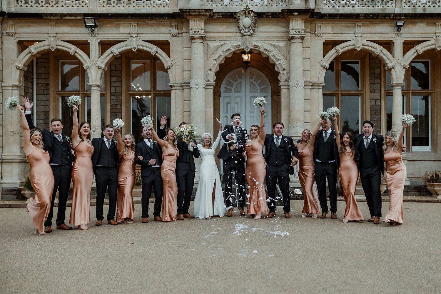 Wedding party in coordinated attire poses together outside a beautiful stone mansion.