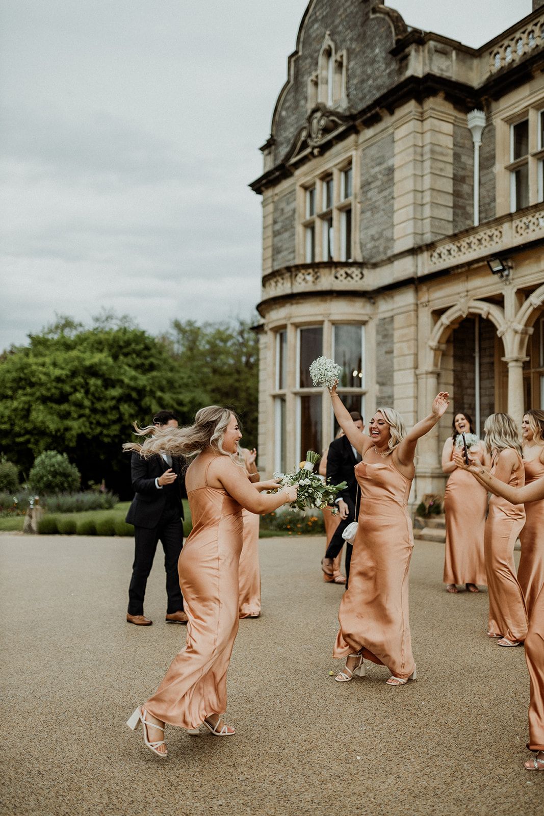 Bridesmaids in flowing peach gowns dance and twirl on the grounds of an elegant stone mansion.