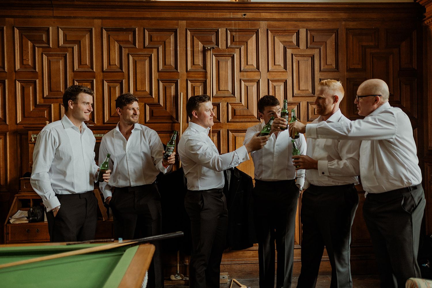 A group in white shirts celebrate with a toast near a pool table in a room with decorative wooden wall paneling.