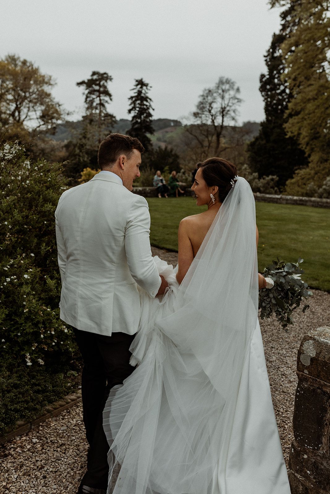 A bride and groom walk together along a rocky path surrounded by autumn trees and natural scenery.