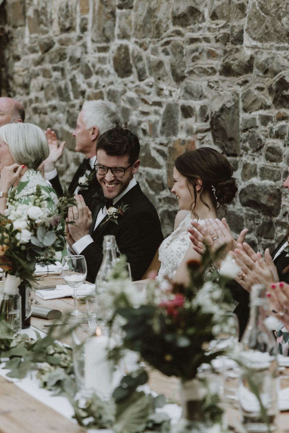Wedding guests applauding during reception dinner at stone-walled venue with floral centerpieces.