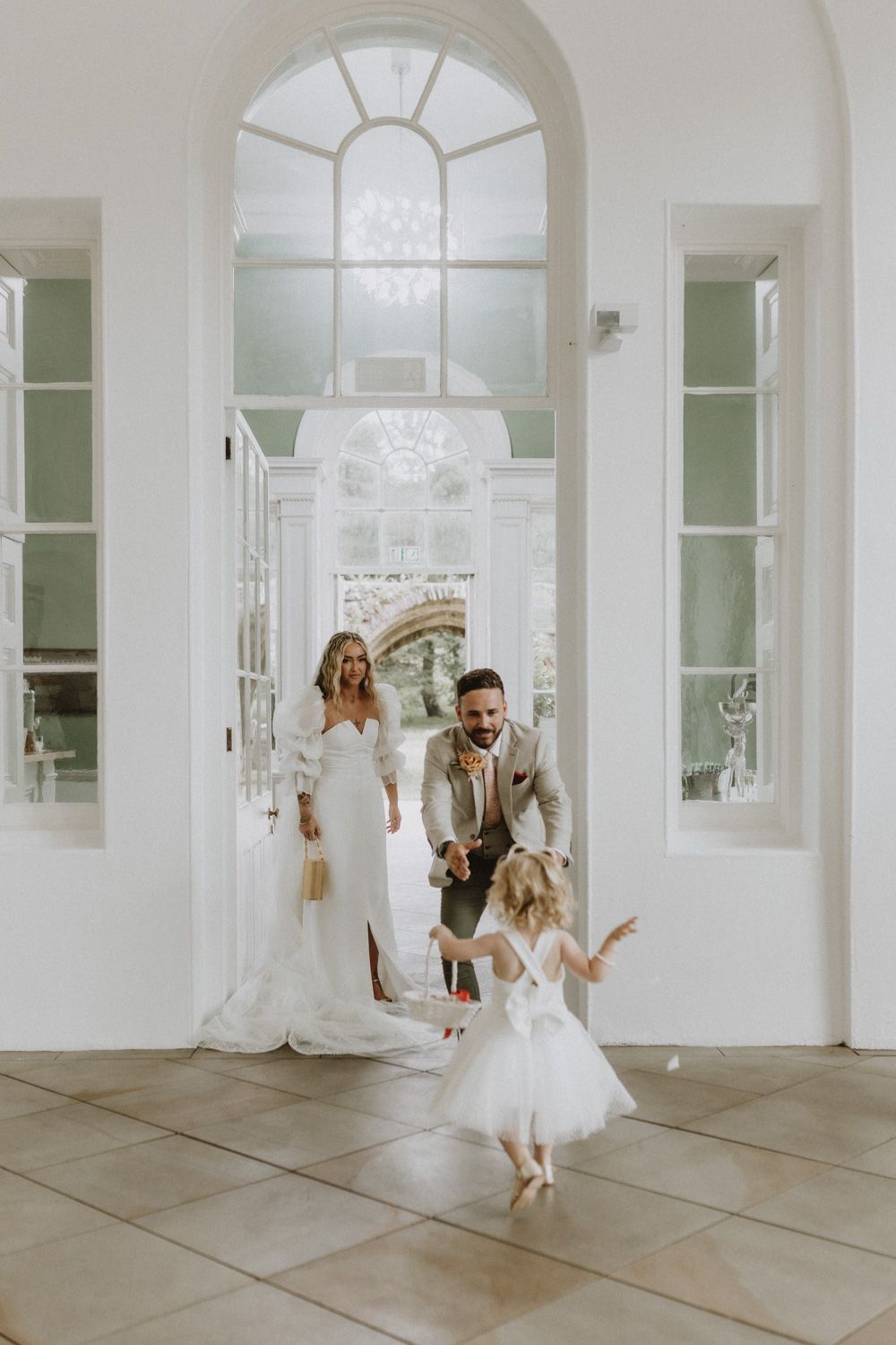 Little girl in white dress dances joyfully during wedding celebration in bright interior with arched windows.