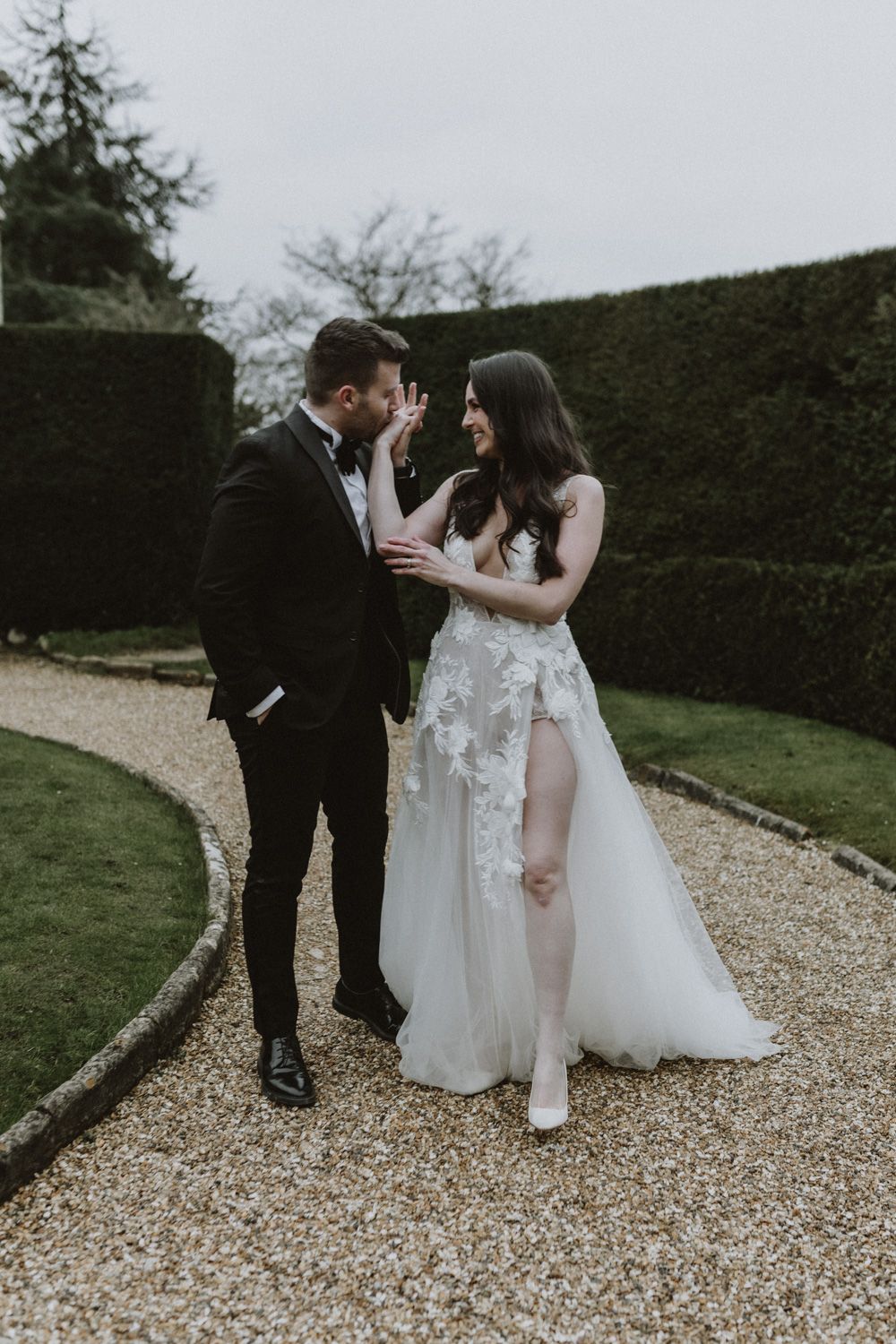 A tender moment shared on a gravel path with manicured hedges creating an elegant backdrop.