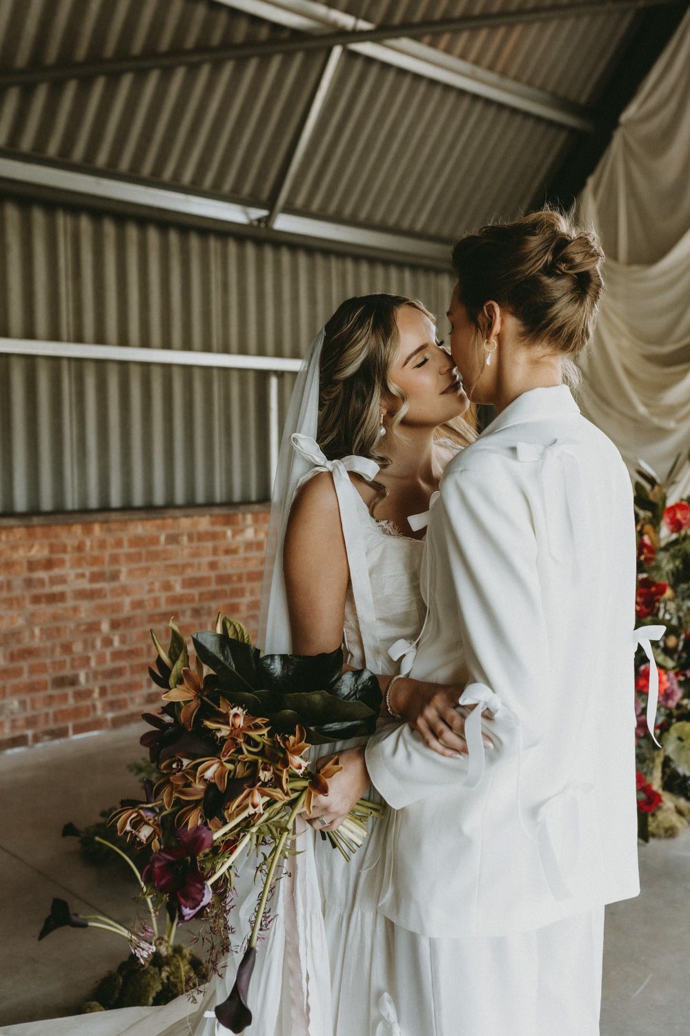 Romantic embrace between two figures in white against an industrial brick wall setting.