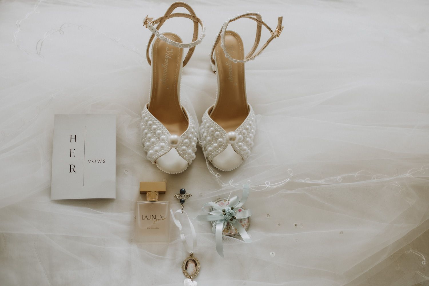 Elegant bridal accessories including gold shoes, perfume bottle, and delicate white fabric laid out on light surface.