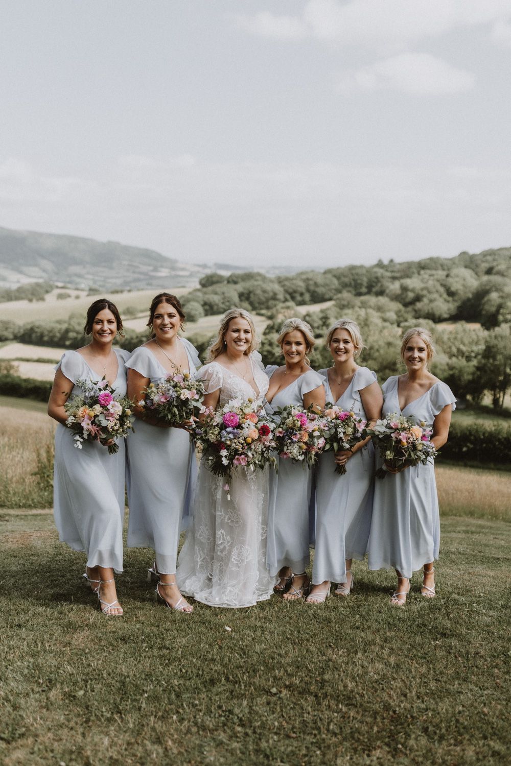 Bridesmaids in pale blue dresses pose with bouquets against a scenic countryside backdrop.
