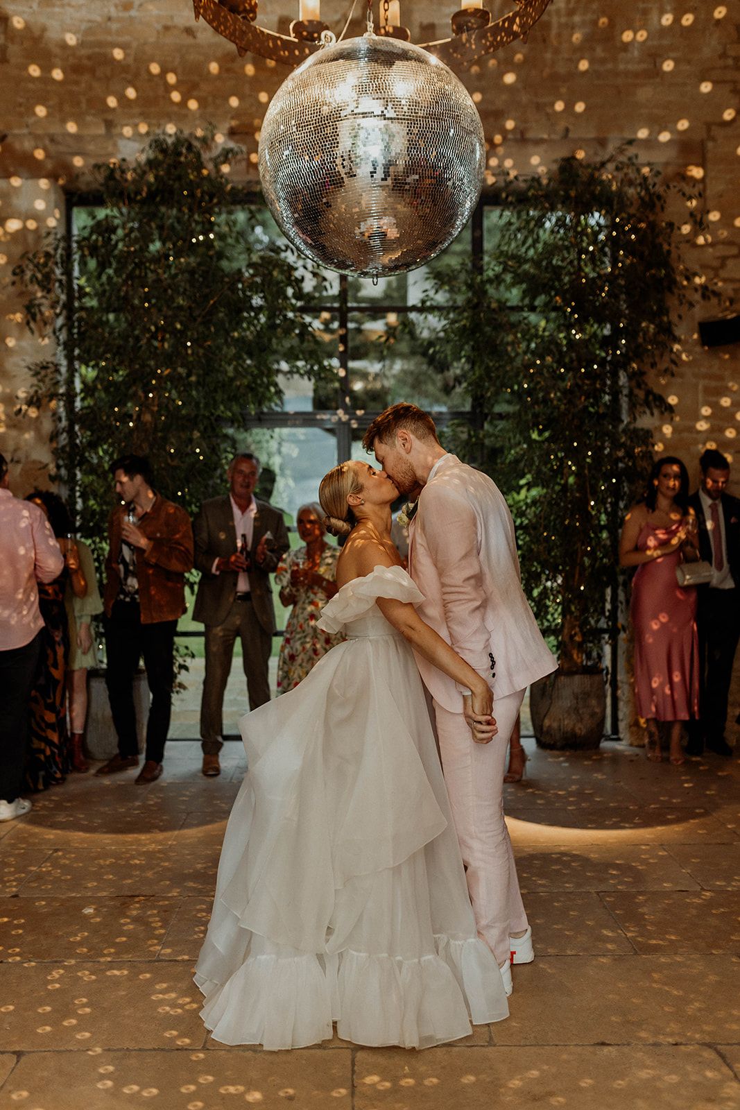 A couple dances under a sparkling disco ball surrounded by greenery and soft lighting at their wedding reception.