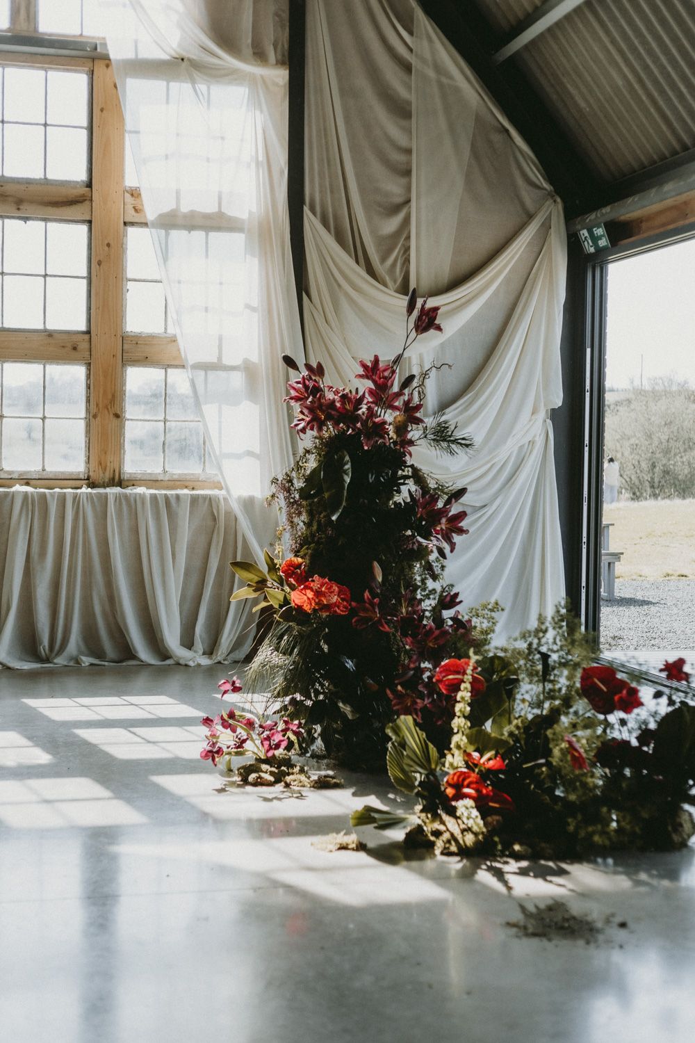 Elegant floral arrangements with red blooms decorate a corner against white draped fabric.