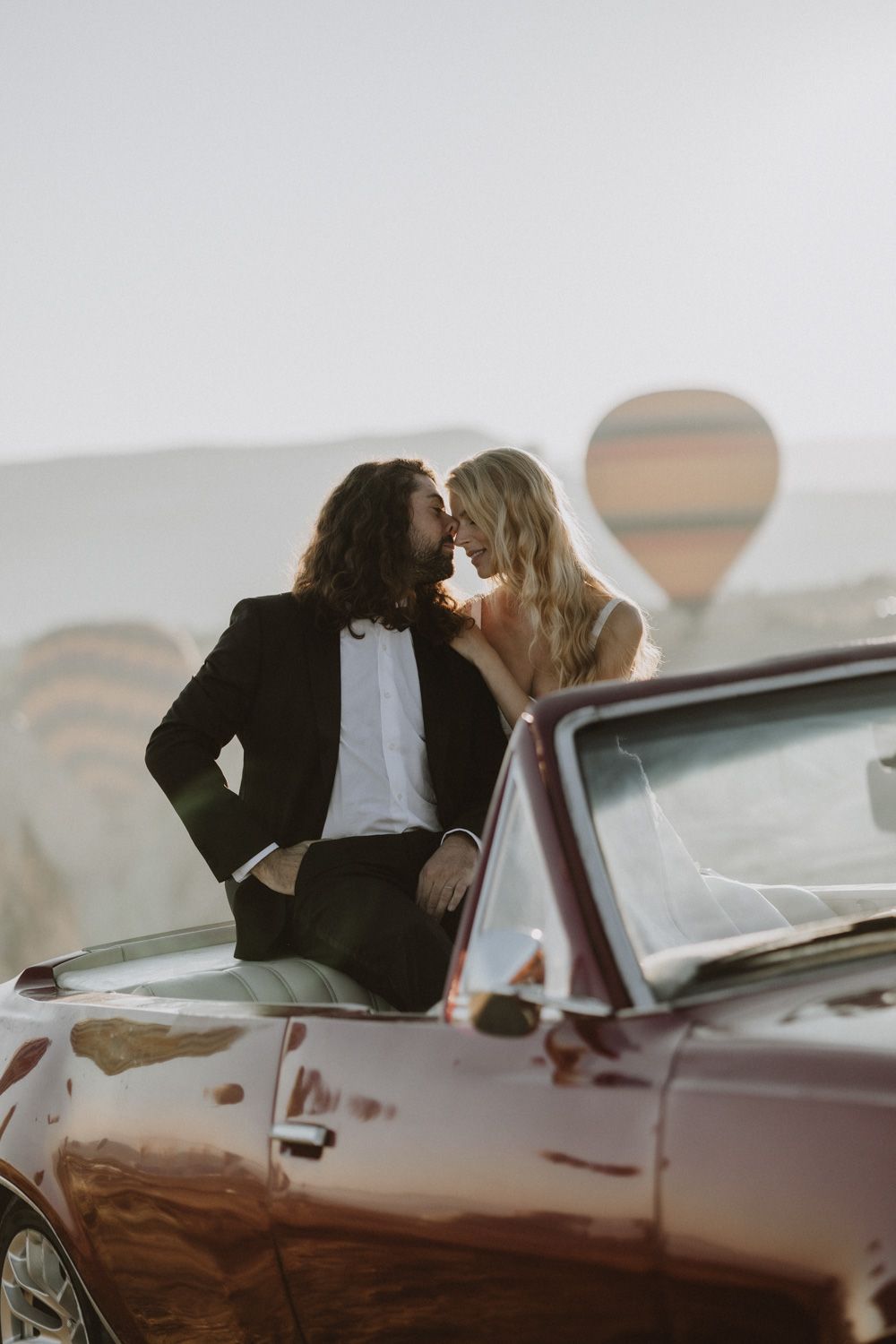 Romantic moment shared between newlyweds leaning against a vintage car at sunset.