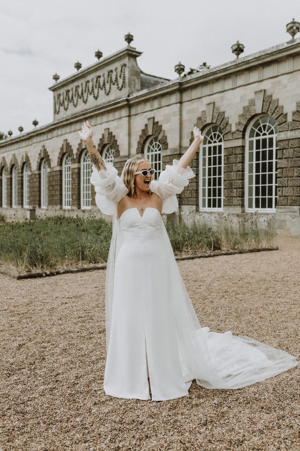 Bride in flowing white dress celebrating with arms raised outside ornate building.