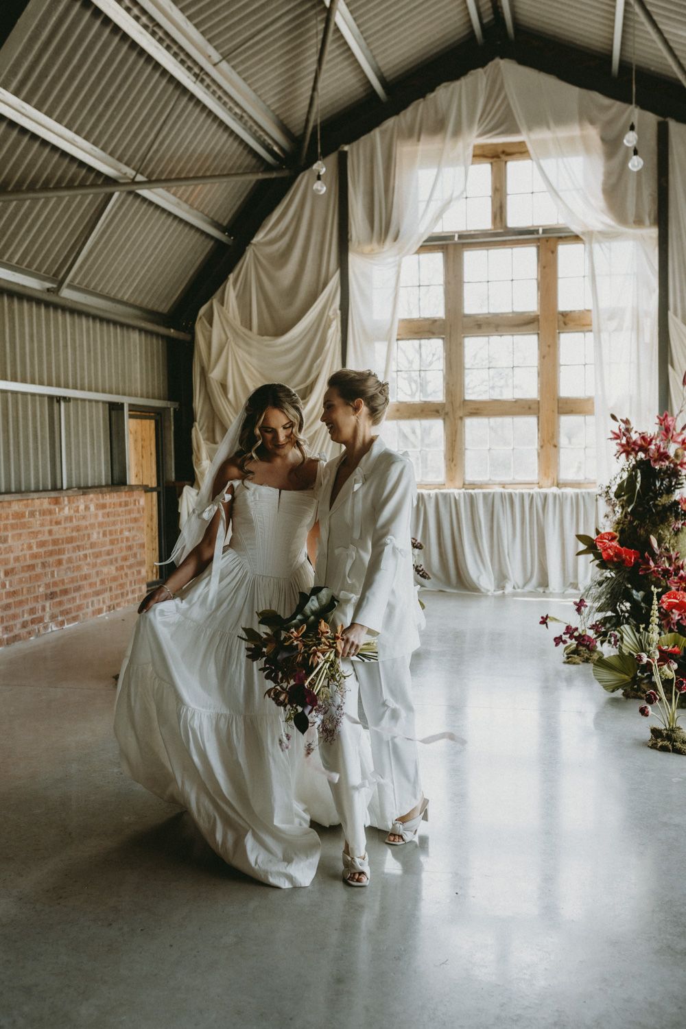 Wedding couple in flowing white attire dancing in sunlit industrial venue with floral arrangements.