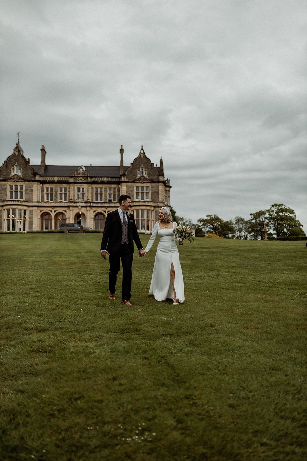 Wedding couple strolls across green lawn towards historic Gothic Revival mansion under cloudy skies.