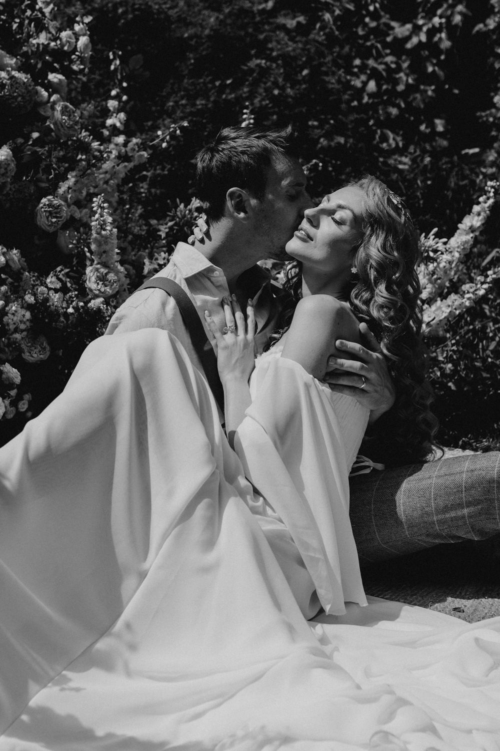 Black and white photo of a couple in elegant white attire sharing an intimate moment outdoors.