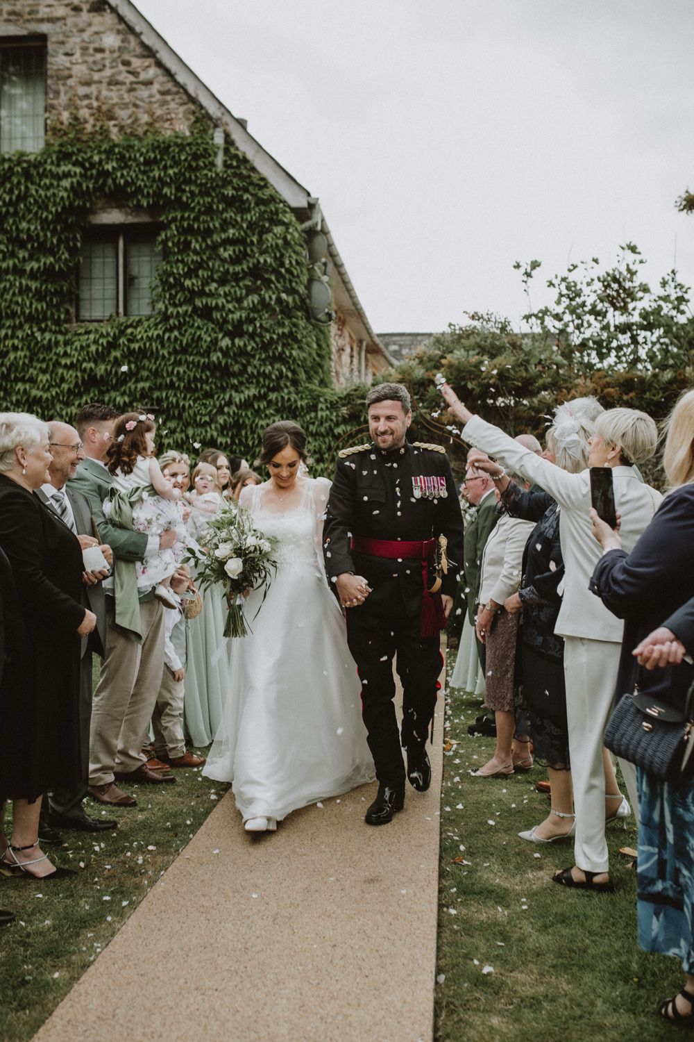 Wedding guests stand in a row throwing confetti over a bride and groom as they exit their ceremony.