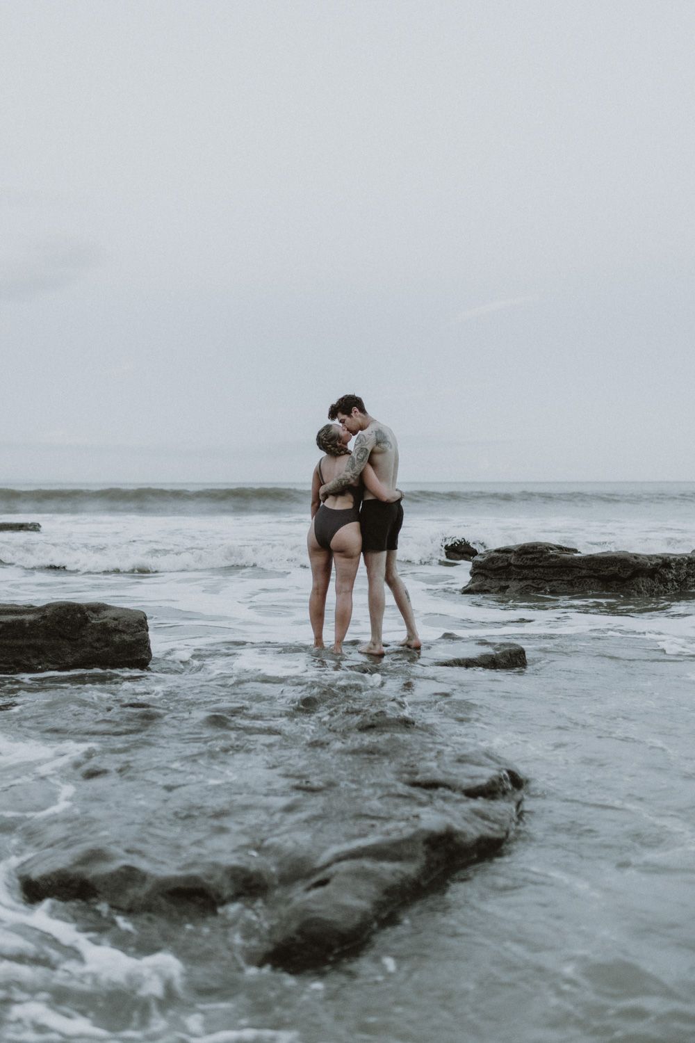 Intimate couple embrace on rocky coastline with misty ocean backdrop.