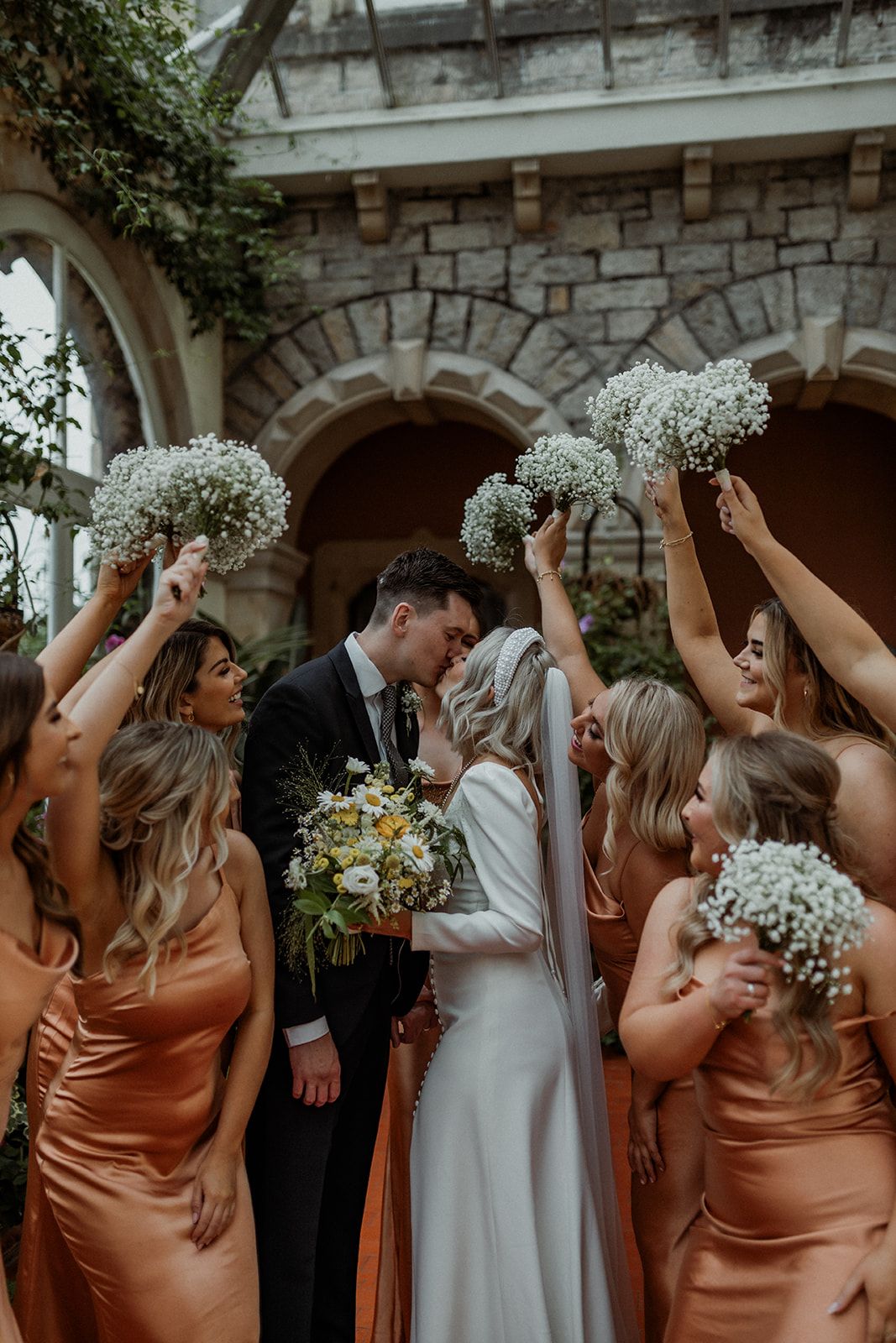 Wedding party in copper-colored bridesmaid dresses and white gown celebrate with baby's breath bouquets at stone venue.