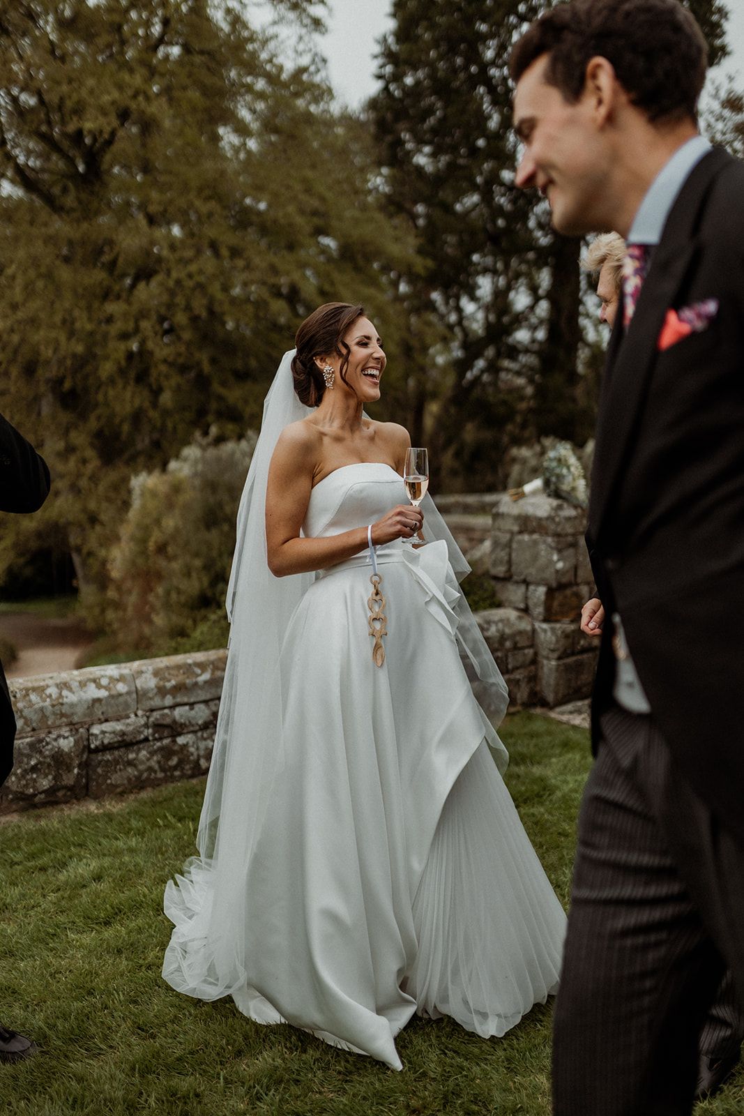 Happy bride in flowing white wedding dress shares joyful moment with groom on a grassy lawn near stone wall.