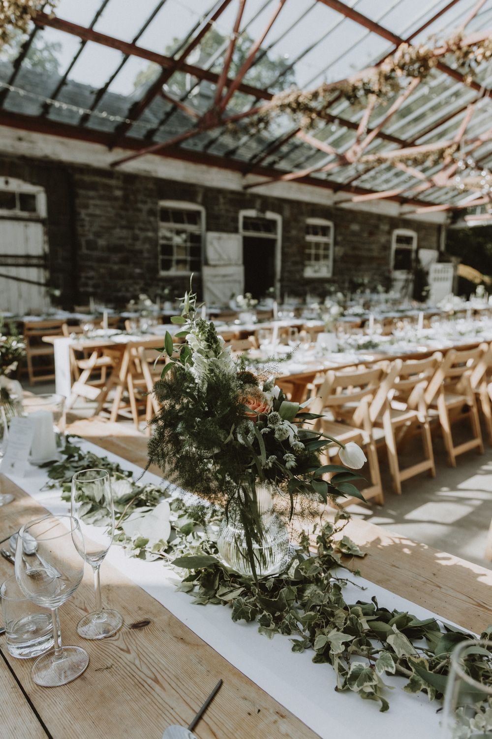 Rustic wedding reception setup with long tables adorned with greenery garlands in a greenhouse venue.