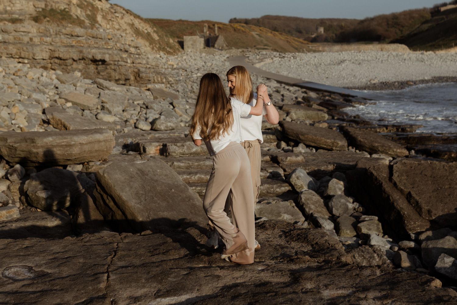 Couple embraces on rocky coastal cliffs at sunset with dramatic ocean and cliff backdrop.
