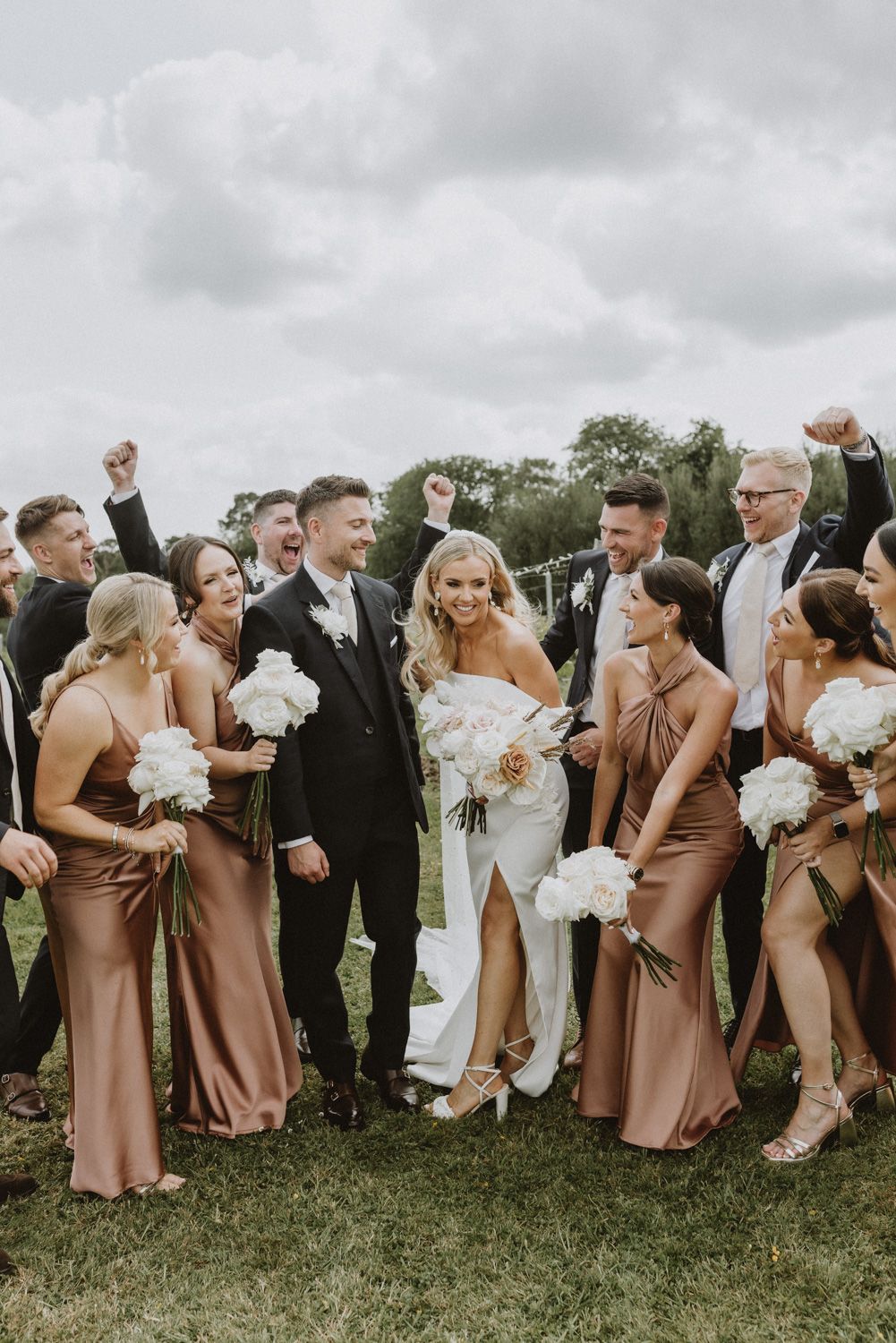 Wedding party in brown bridesmaid dresses and black suits pose together on grassy field.