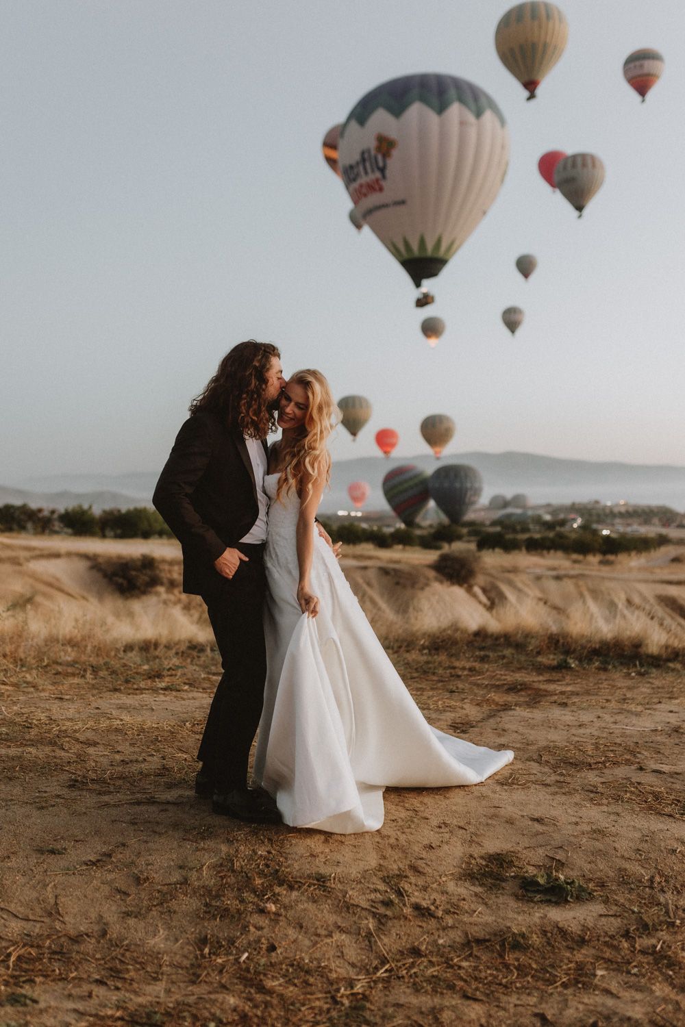 Elegant wedding couple standing on hilltop with colorful hot air balloons floating in background during sunset.