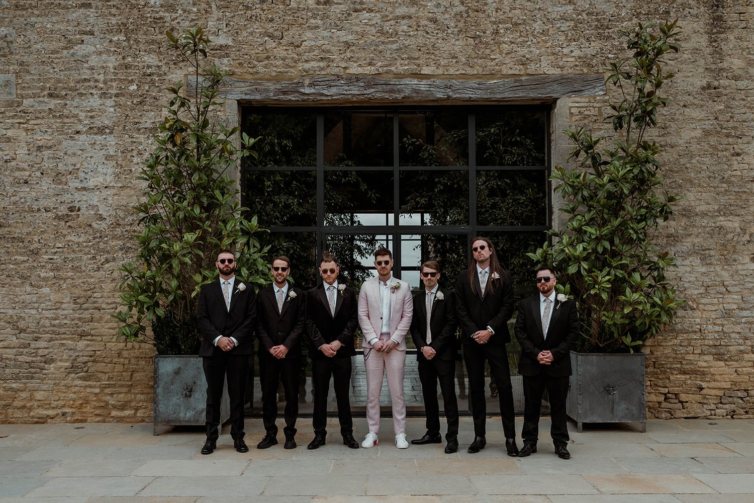 A wedding party group poses in front of a rustic stone wall during an outdoor celebration.