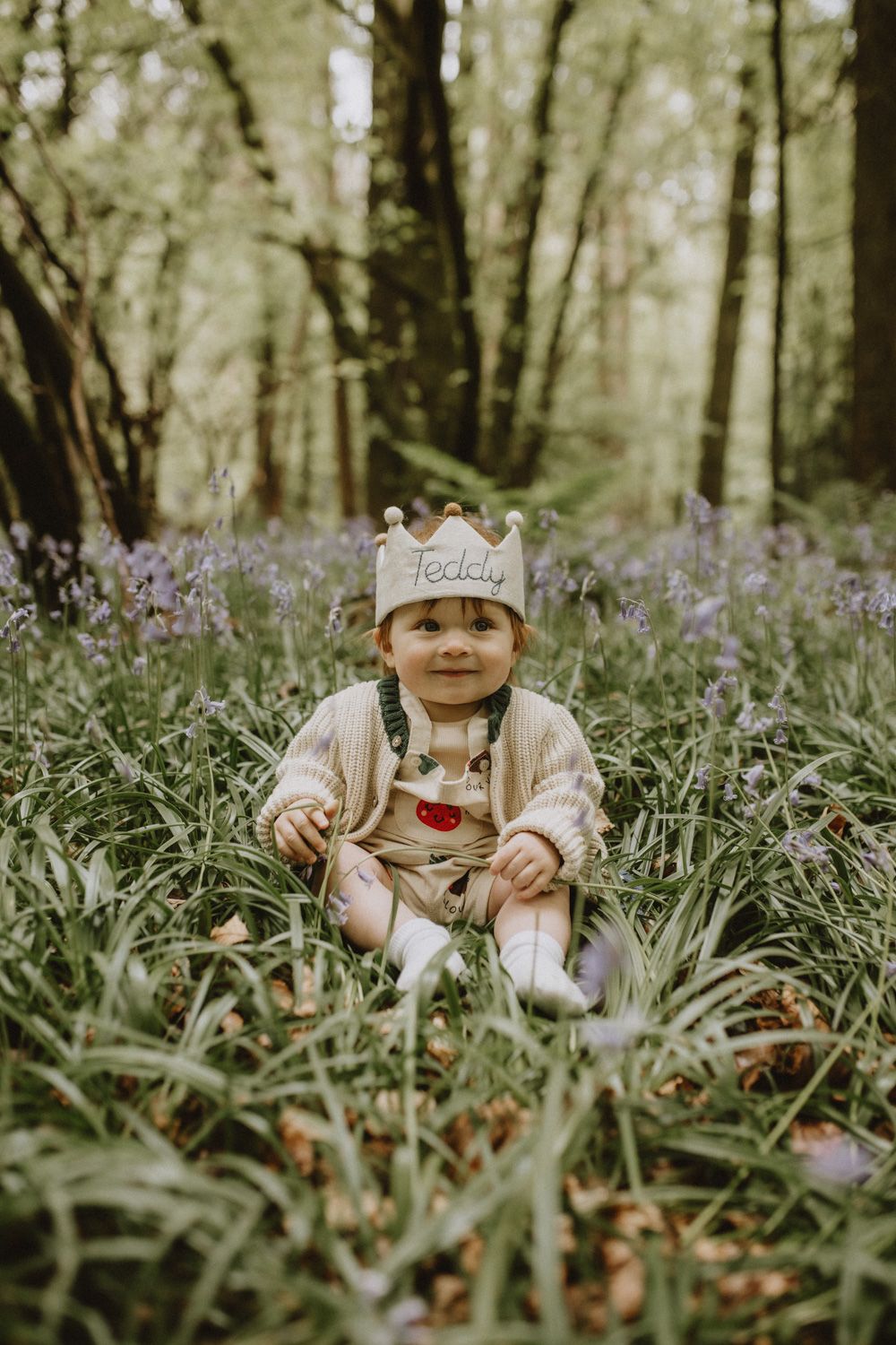 Baby wearing a cap sits among purple wildflowers in a forest setting.