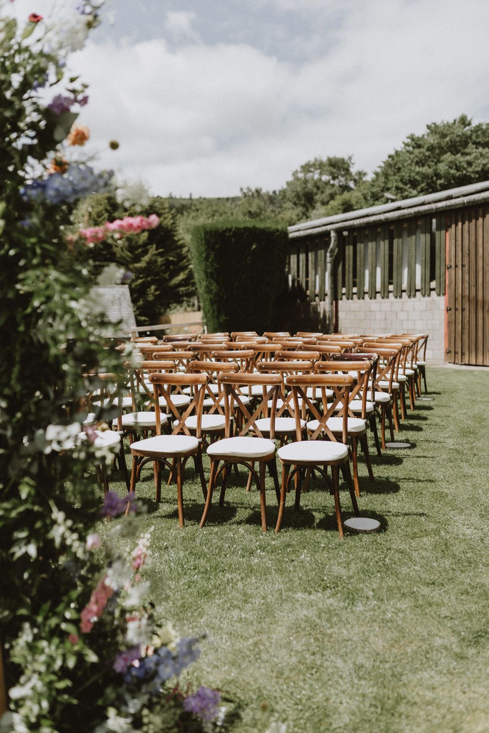 Outdoor wedding ceremony setup with wooden cross-back chairs arranged on a green lawn.