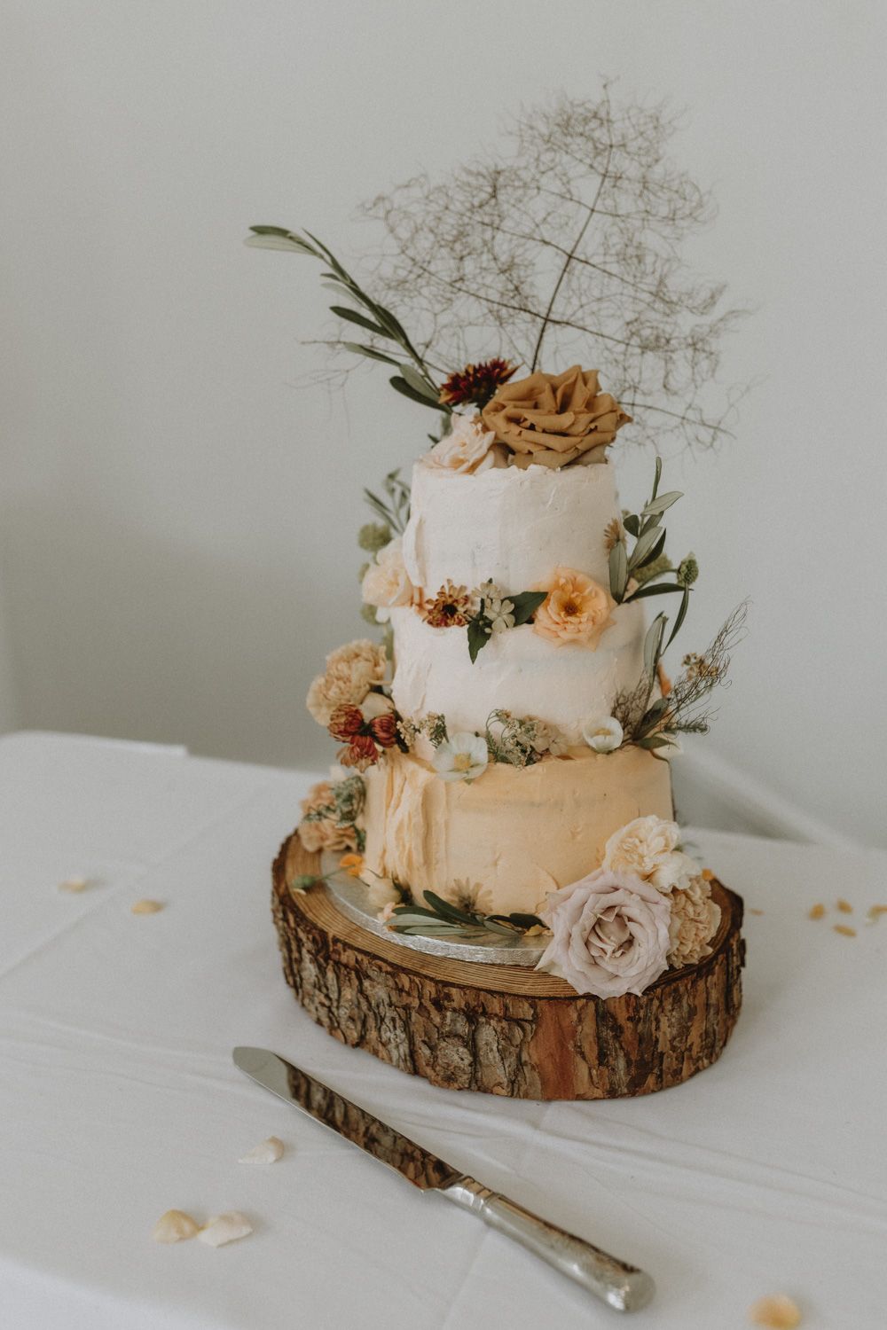 Rustic three-tier wedding cake decorated with dried flowers and roses on a wooden slice base.