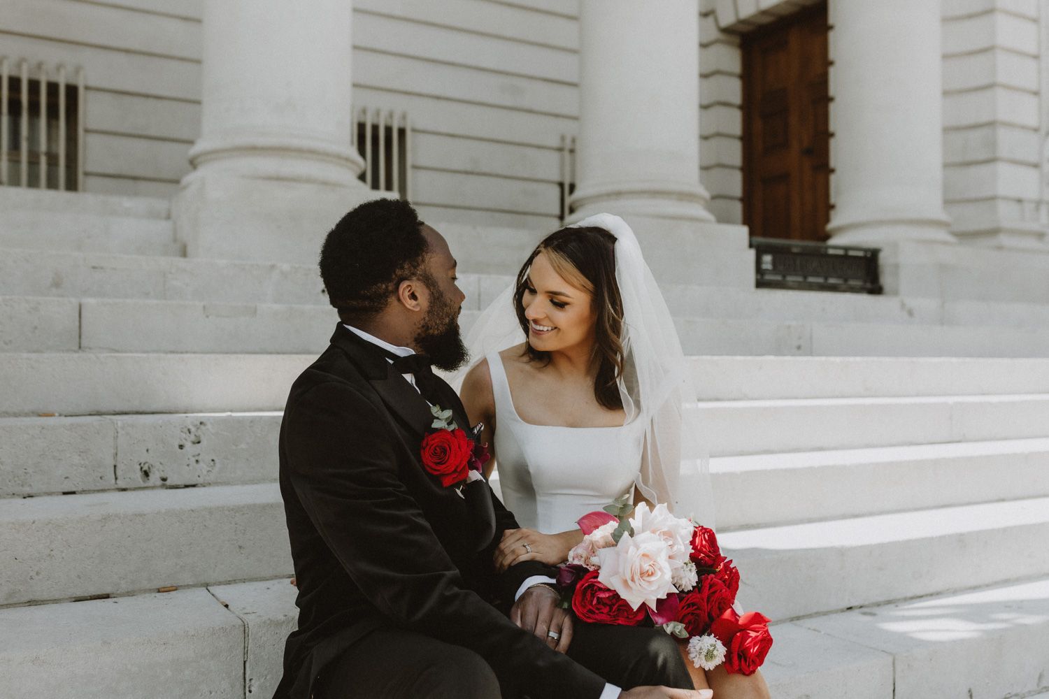 Newlyweds share romantic moment on white stone steps with red bouquet.