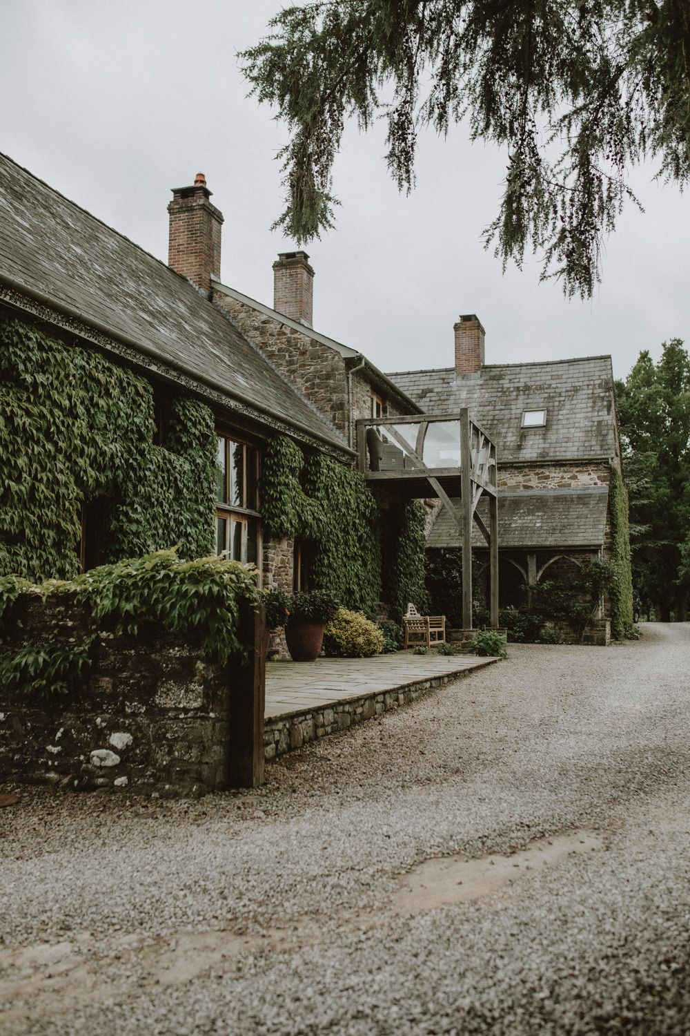 An ivy-covered stone cottage with multiple chimneys and a gravel driveway set against an overcast sky.