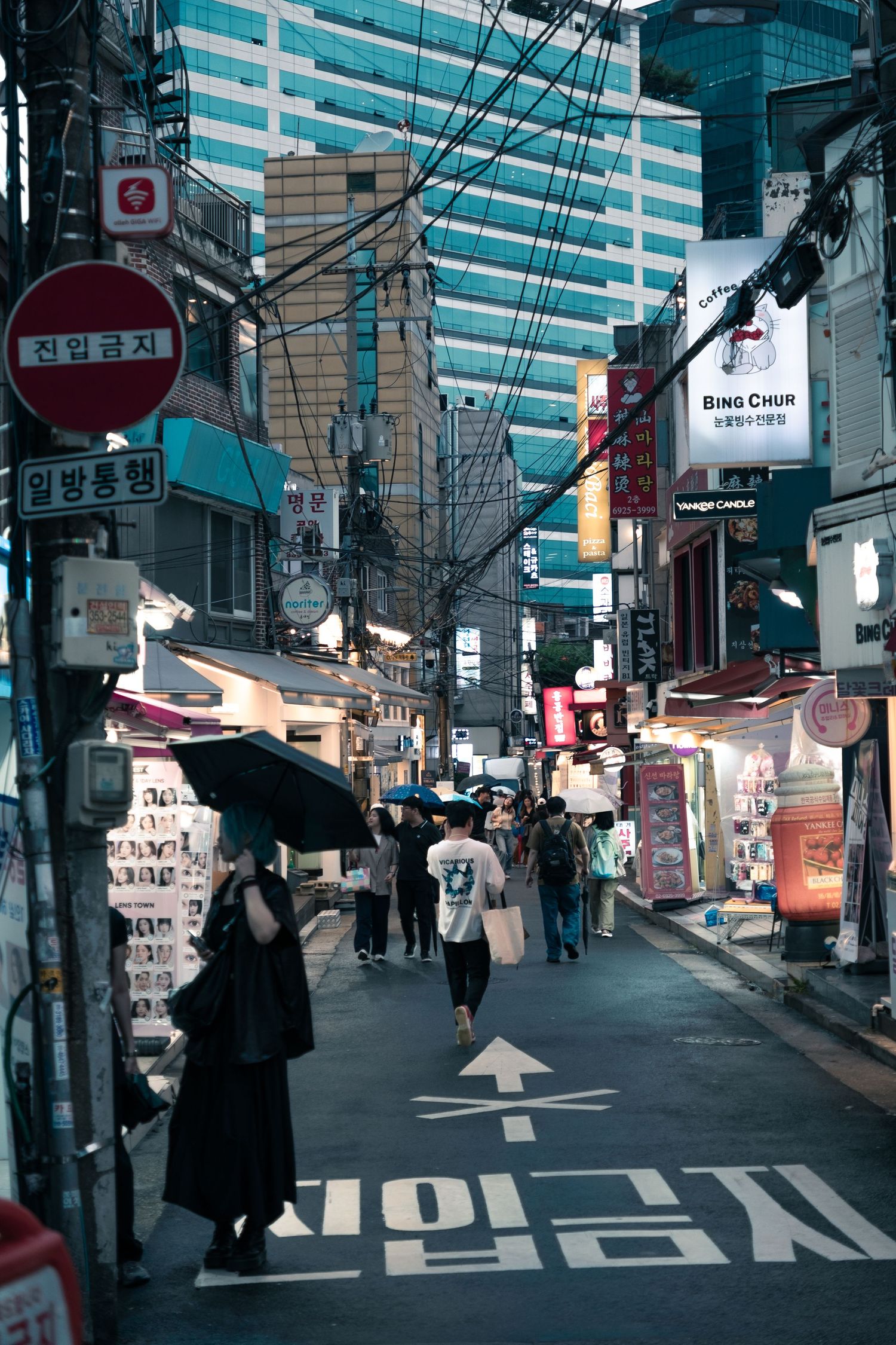 Urban street scene in South Korea with modern buildings, signage, and pedestrians walking under power lines at dusk.