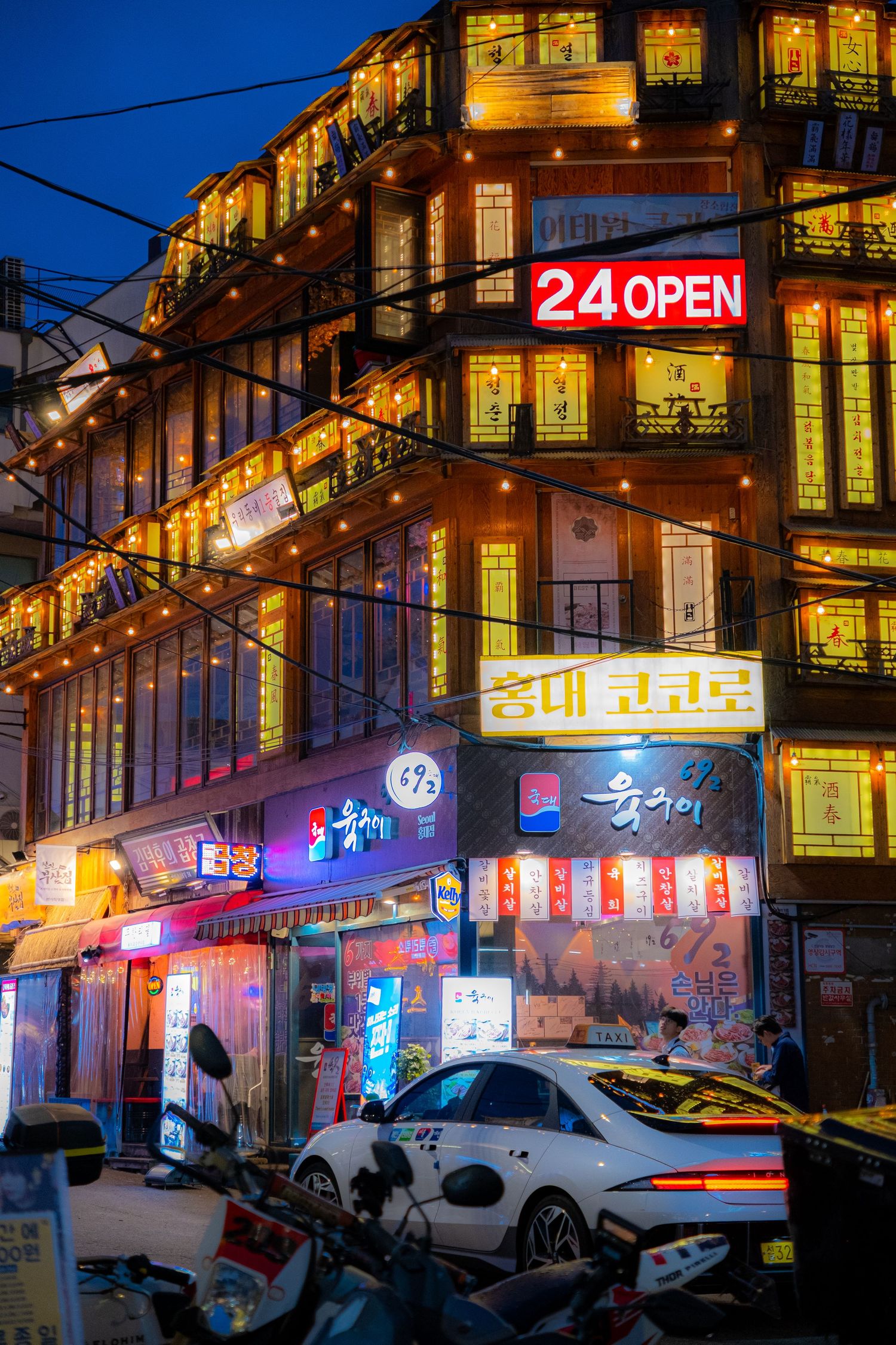 Illuminated Korean street scene at night with neon signs, multi-story building and parked cars on a busy urban street.