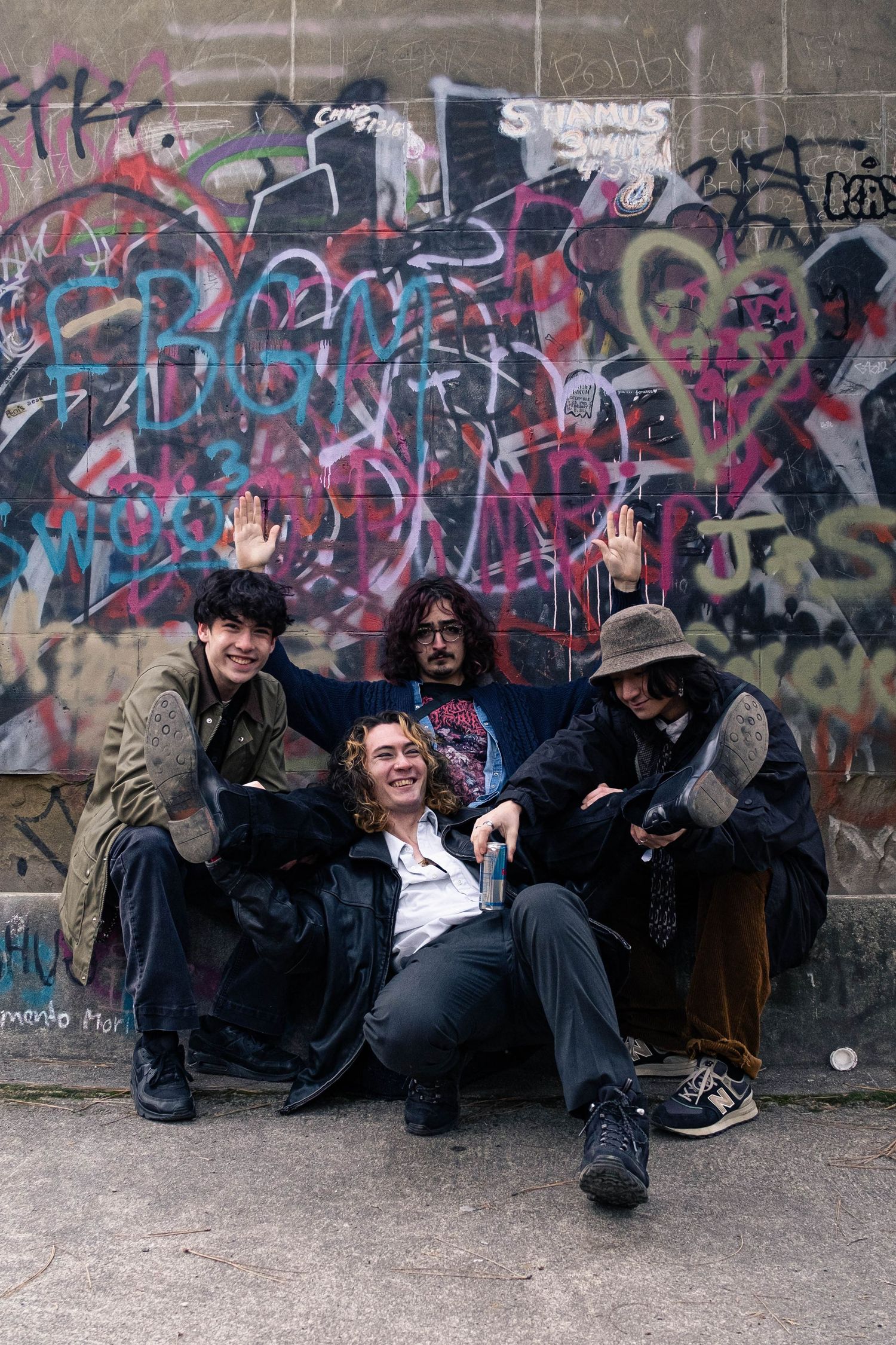 Group of people sitting casually against graffiti wall in urban setting wearing dark clothing.