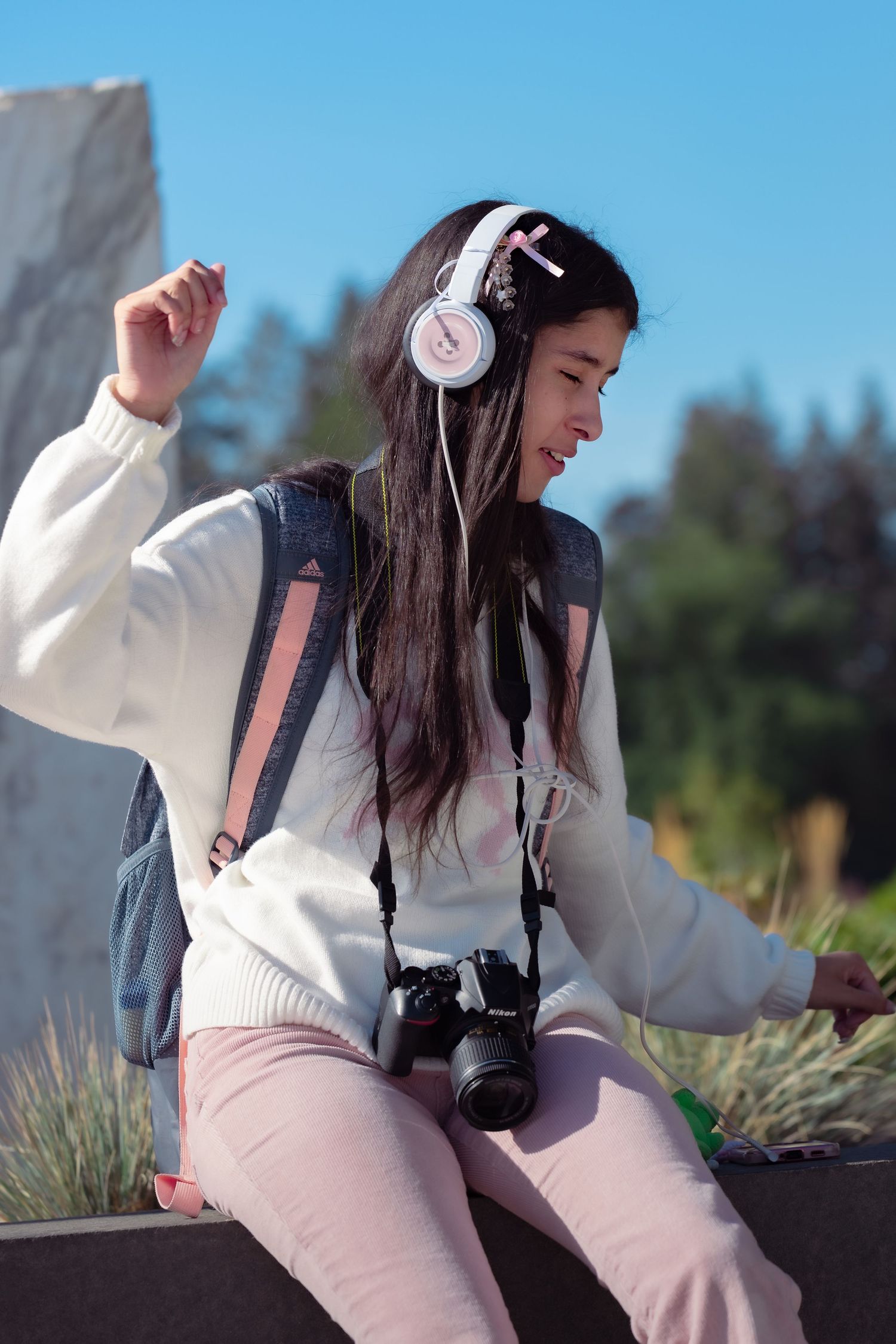 A photographer in a white sweater and pink pants sits outdoors with a camera around their neck enjoying a sunny day.