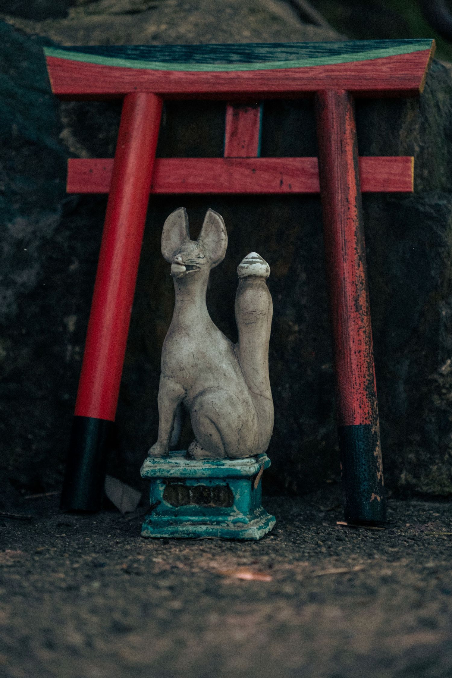 A stone kitsune fox statue sits beneath a miniature red torii gate at a Japanese shrine.