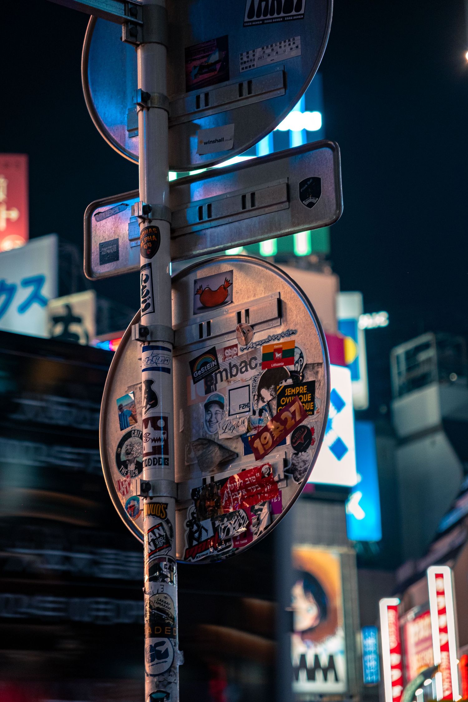 A street sign covered in stickers at night with colorful neon lights and urban reflections in Tokyo's city streets.