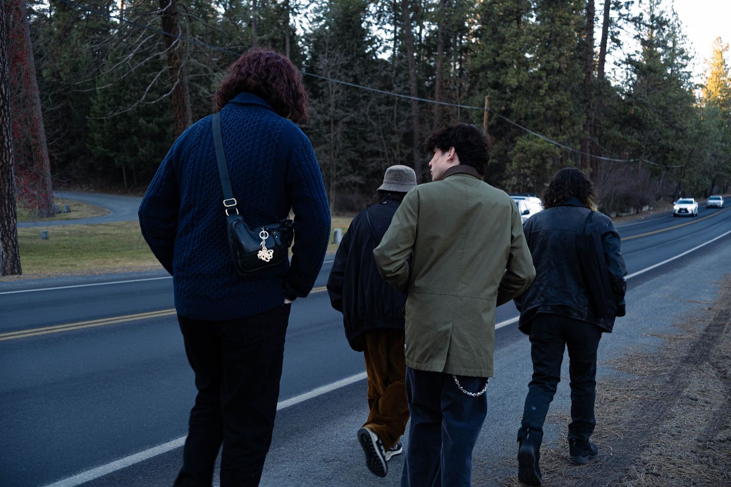 A group of people walking along a dark wooded road at dusk with trees visible in the background.