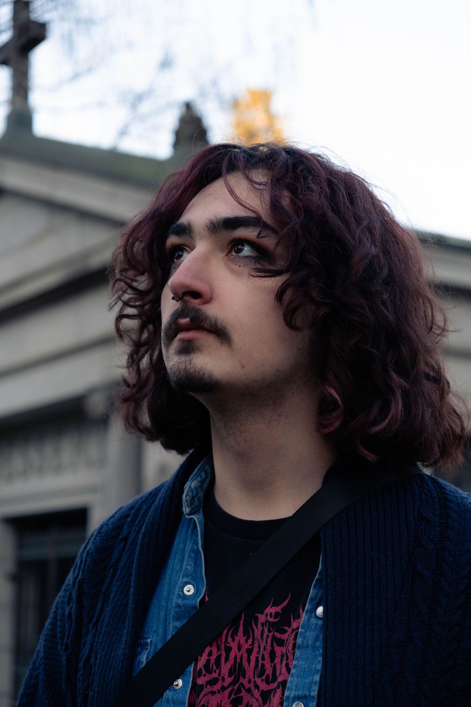 Person with dark curly hair and mustache wearing a blue jacket and burgundy shirt looking upward against an urban backdrop.