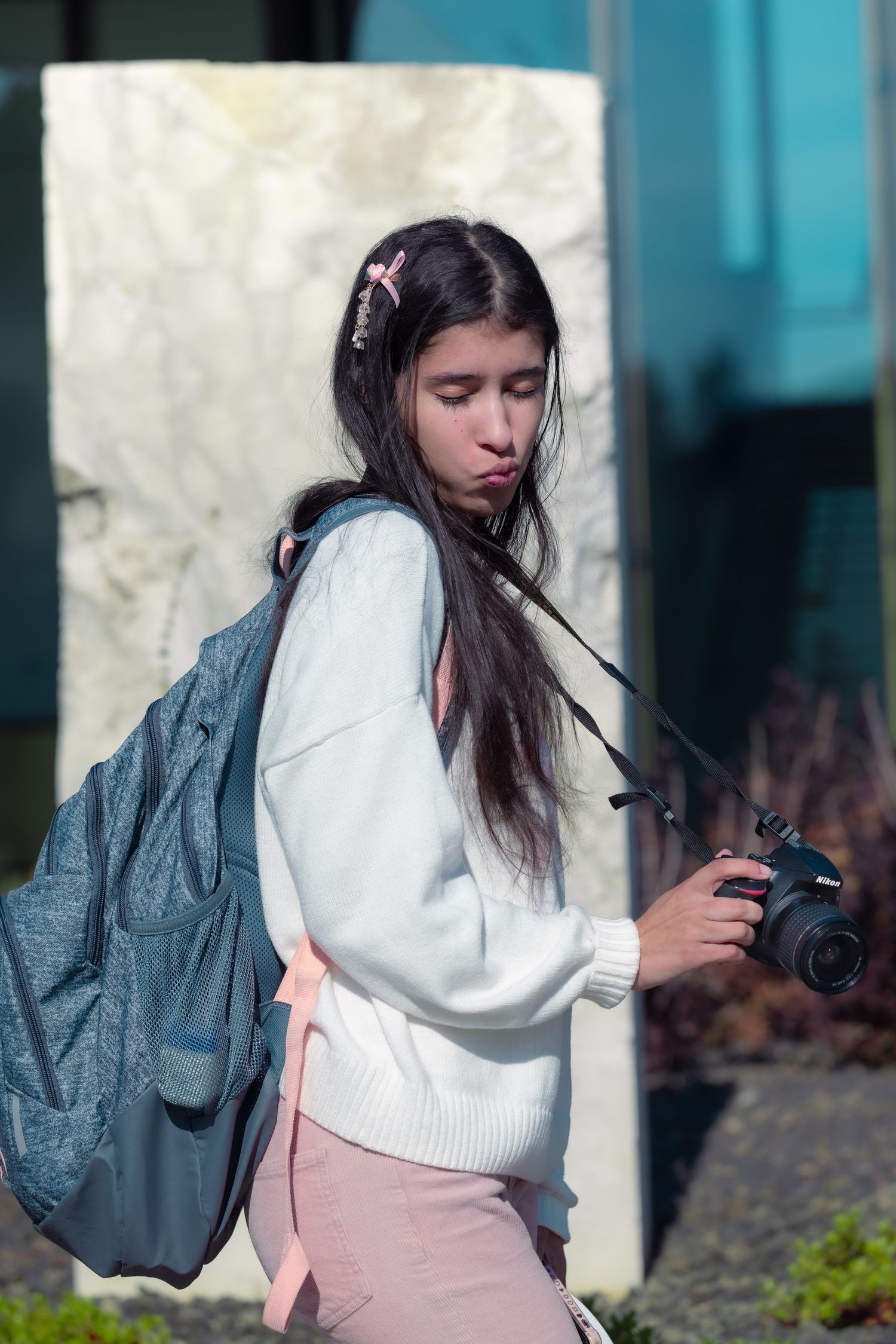 A student with long dark hair wearing a white sweater and backpack holds a camera while standing outdoors.