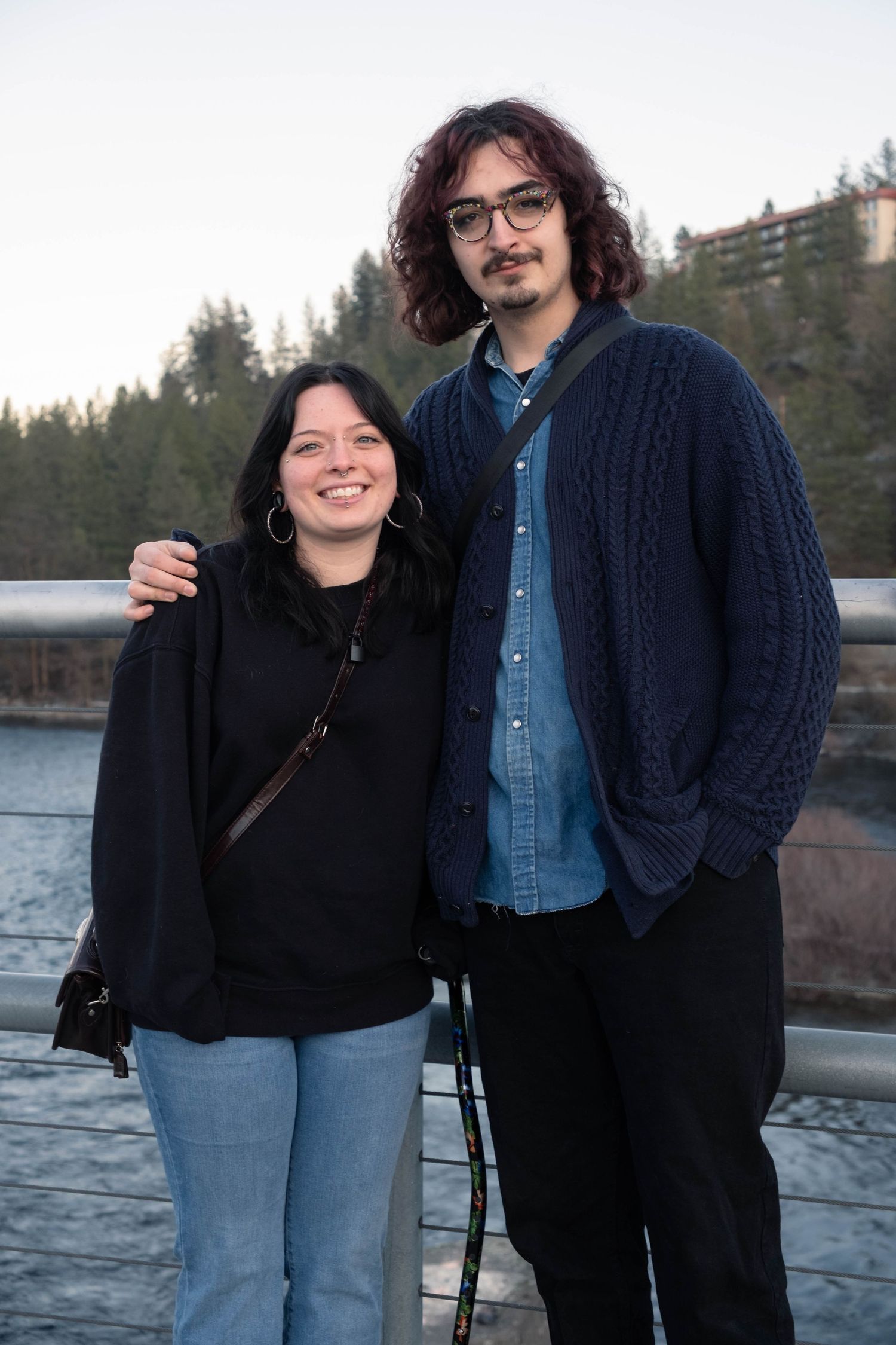 Two people stand together outdoors near a lake with mountains and trees visible in the background.