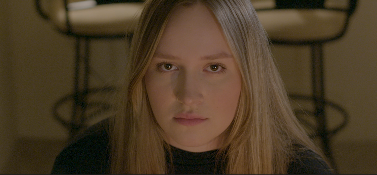 Person with long light hair sitting in muted lighting with contemplative expression.