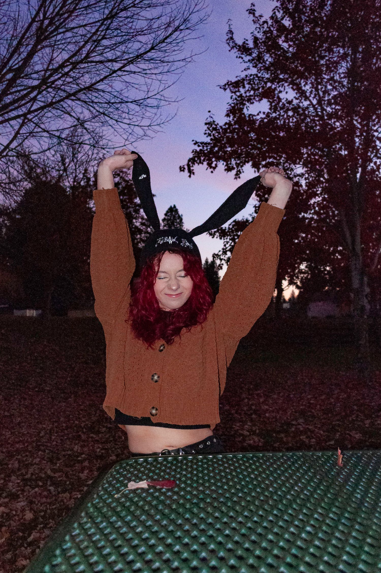 Person in brown sweater poses playfully on green polka dot surface at dusk with autumn trees in background.