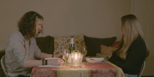 A couple sits together around a dimly lit apartment table, conversing over dinner.