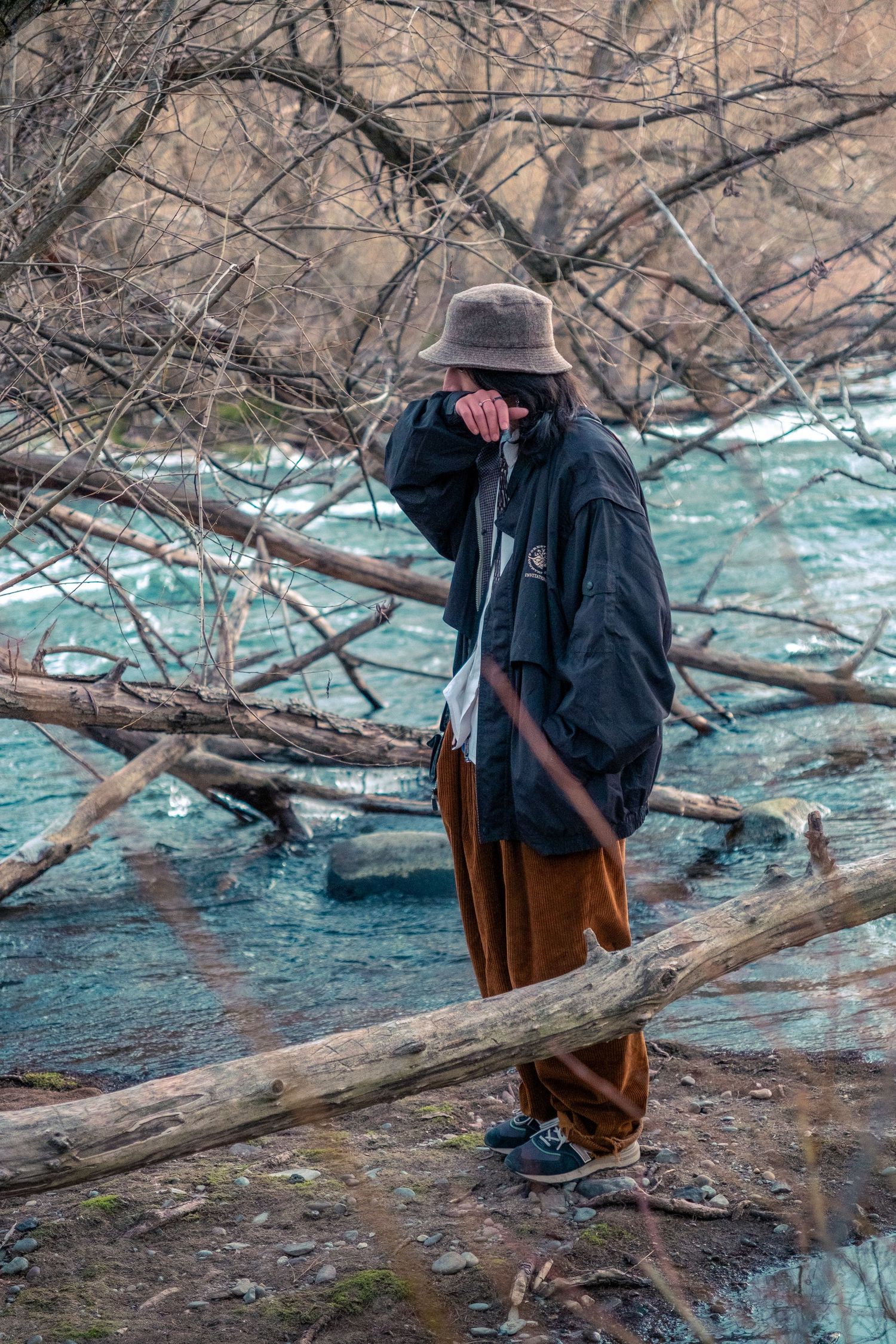 Person in bucket hat and black jacket standing among bare branches near turquoise river.