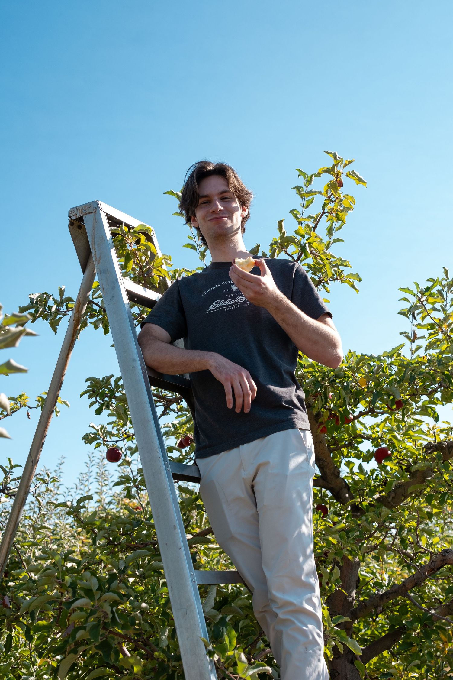 Someone in a dark shirt and light pants stands casually against a white pole with green foliage in the background.